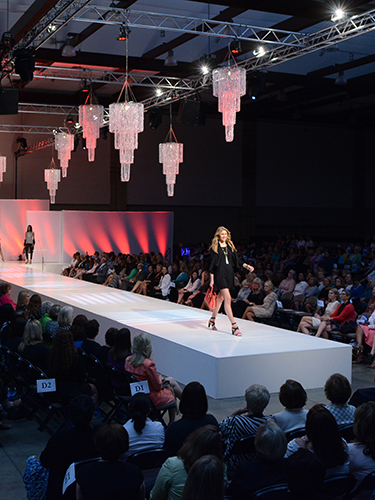 A female model walks down a fashion runway, wearing a stylish summer outfit. The setting features chandeliers hanging above and an audience seated on either side of the stage