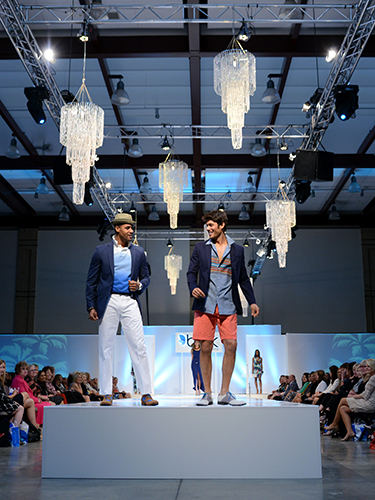 Two male models walk down a fashion runway, wearing stylish summer outfits. The setting features chandeliers hanging above and an audience seated on either side of the stage.