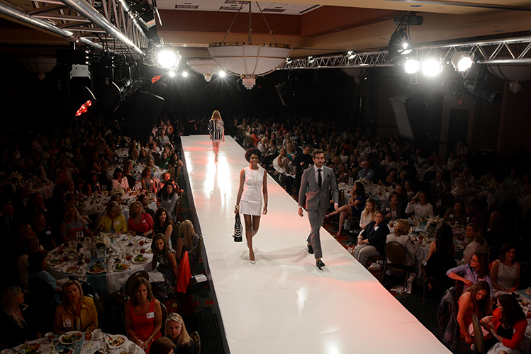 Models walk on a fashion runway with the audience seated at round tables on both sides of the stage. The light rigging from the stage shines bright on the runway.