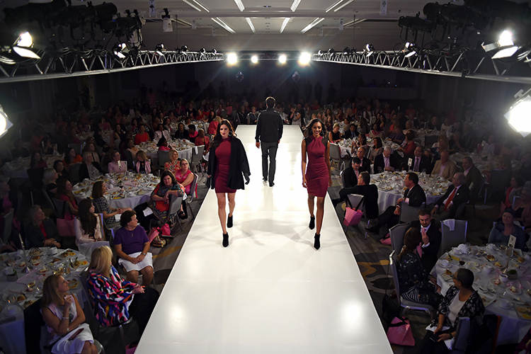 High view of a fashion show scene with three models walking on a brightly lit runway. The audience, seated at round tables, watches the show.