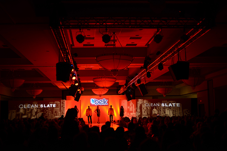 A male and female model walk to the front of a fashion runway dress in stylish clothes. Audience members are seated around the stage.