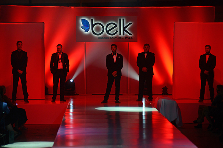 A group of people celebrating on a fashion runway with pink ribbons falling from above. A woman in white holds flowers at center stage, surrounded by others clapping and smiling. A crowd with signs stands behind them in front of a Belk sign