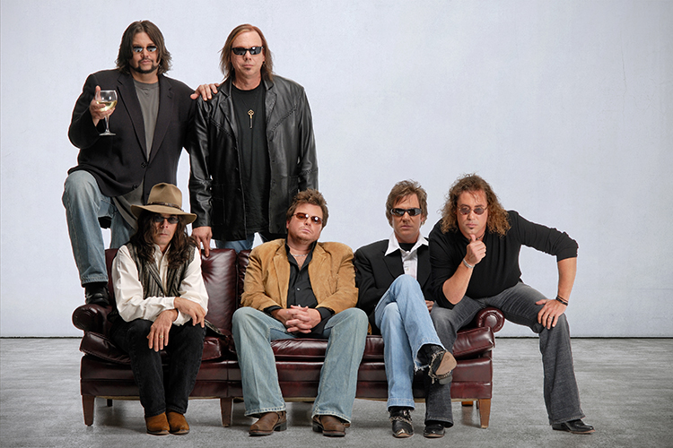 Six men pose for a group photo in casual attire, some wearing sunglasses and leather jackets. They sit and stand around a leather sofa against a plain background.